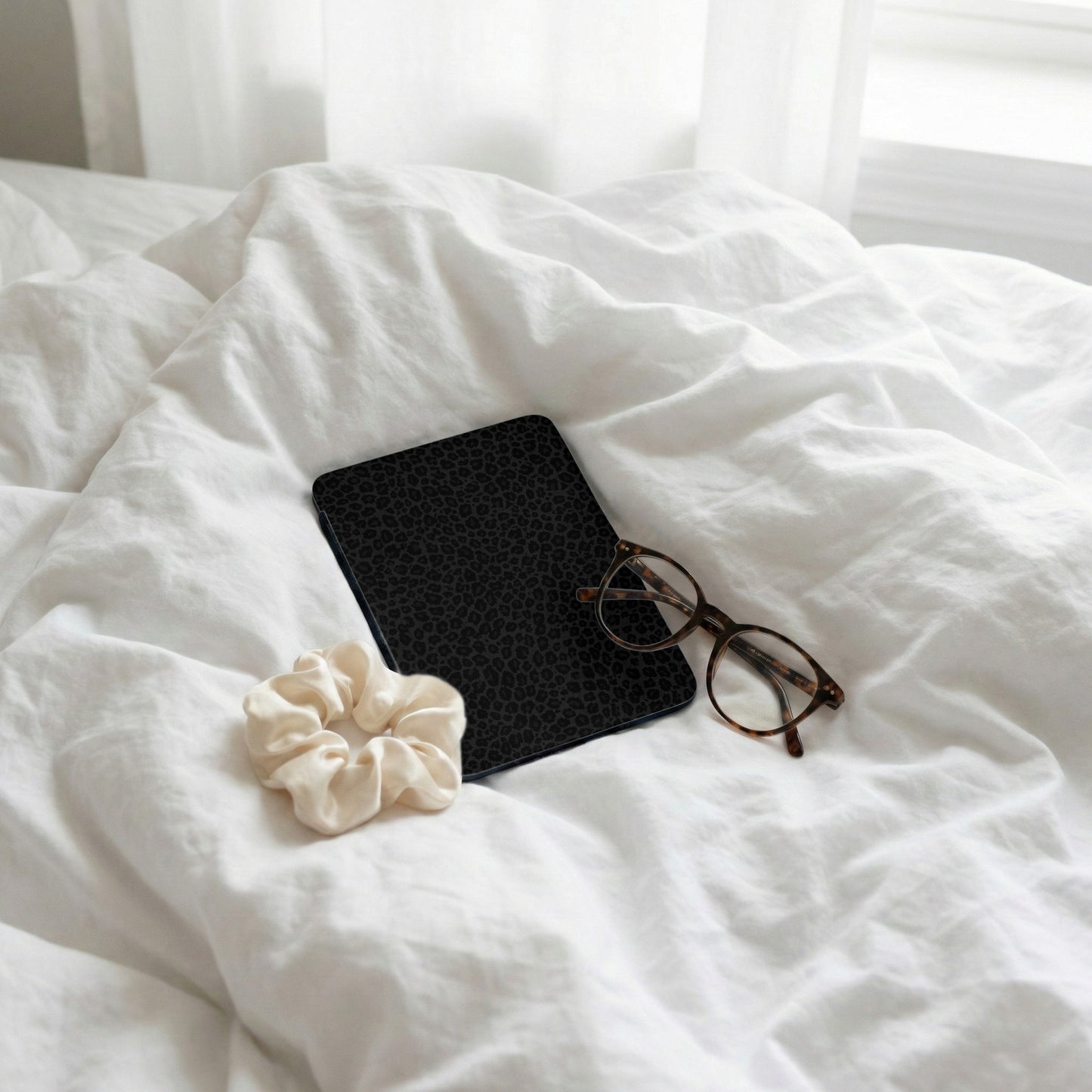 Black leopard print journal with glasses and cream scrunchie on white sheets.