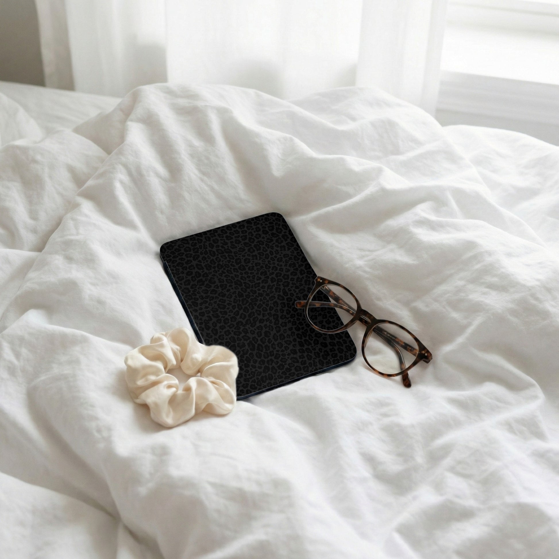 Black leopard print journal with glasses and cream scrunchie on white sheets.