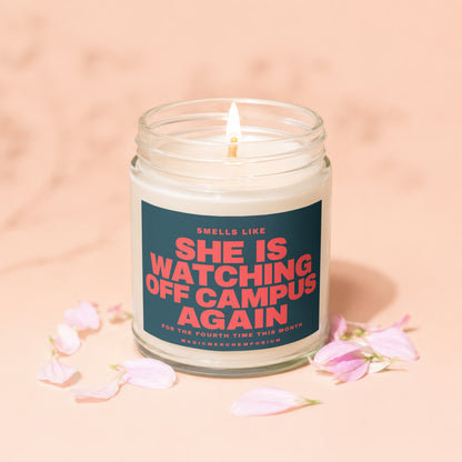 Glass candle with lit wick and label "She is watching off campus again".