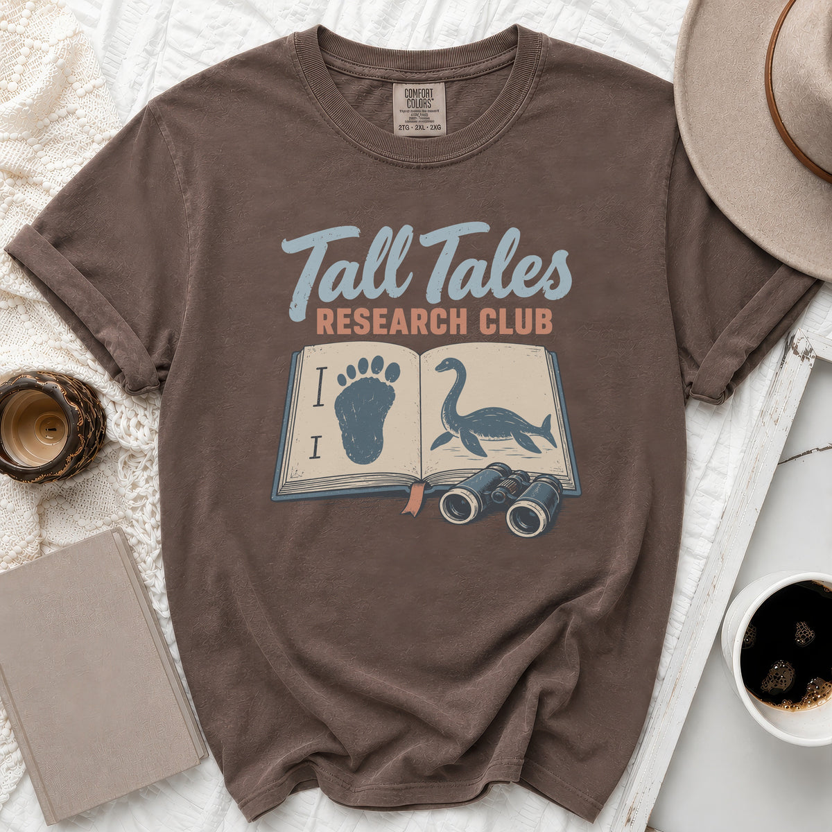 Brown t-shirt with "Tall Tales Research Club" graphic featuring book, Bigfoot footprint, and Loch Ness Monster.