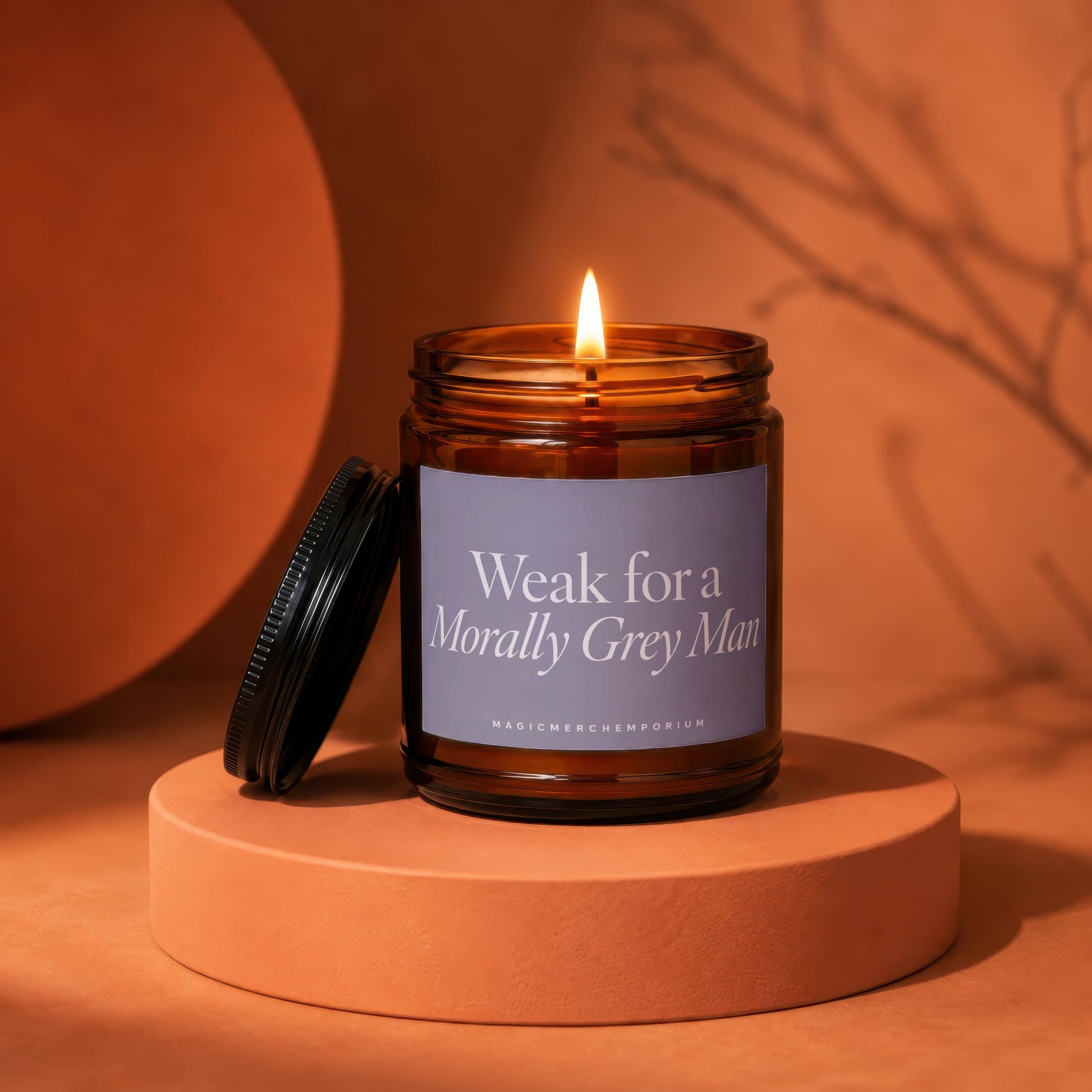 Amber candle with lit wick, lid off, showing label text "Weak for a Morally Grey Man".