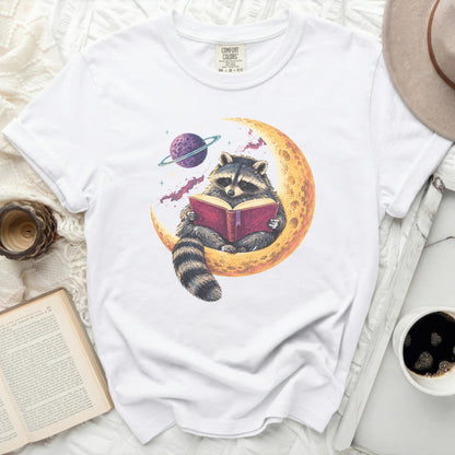 White t-shirt with a graphic of a raccoon reading a book on a crescent moon.