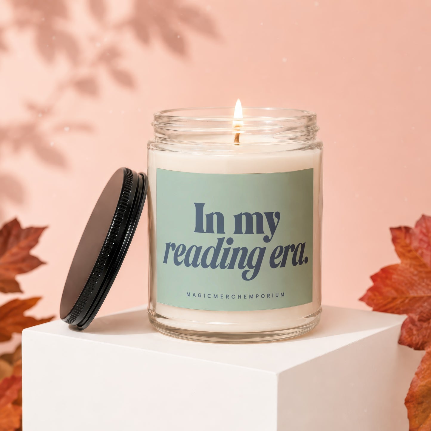 Glass candle with burning wick, lid off, on white cube, text "In my reading era."