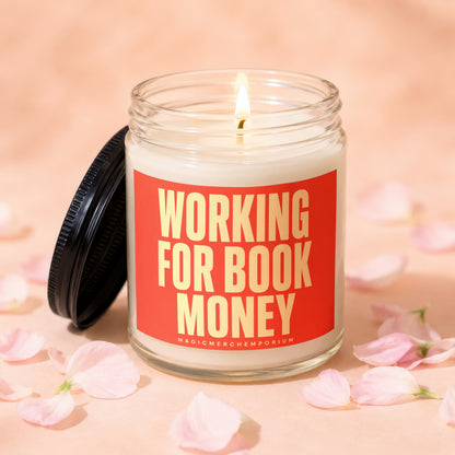 Glass candle with lit wick and "Working for Book Money" text, surrounded by pink petals.