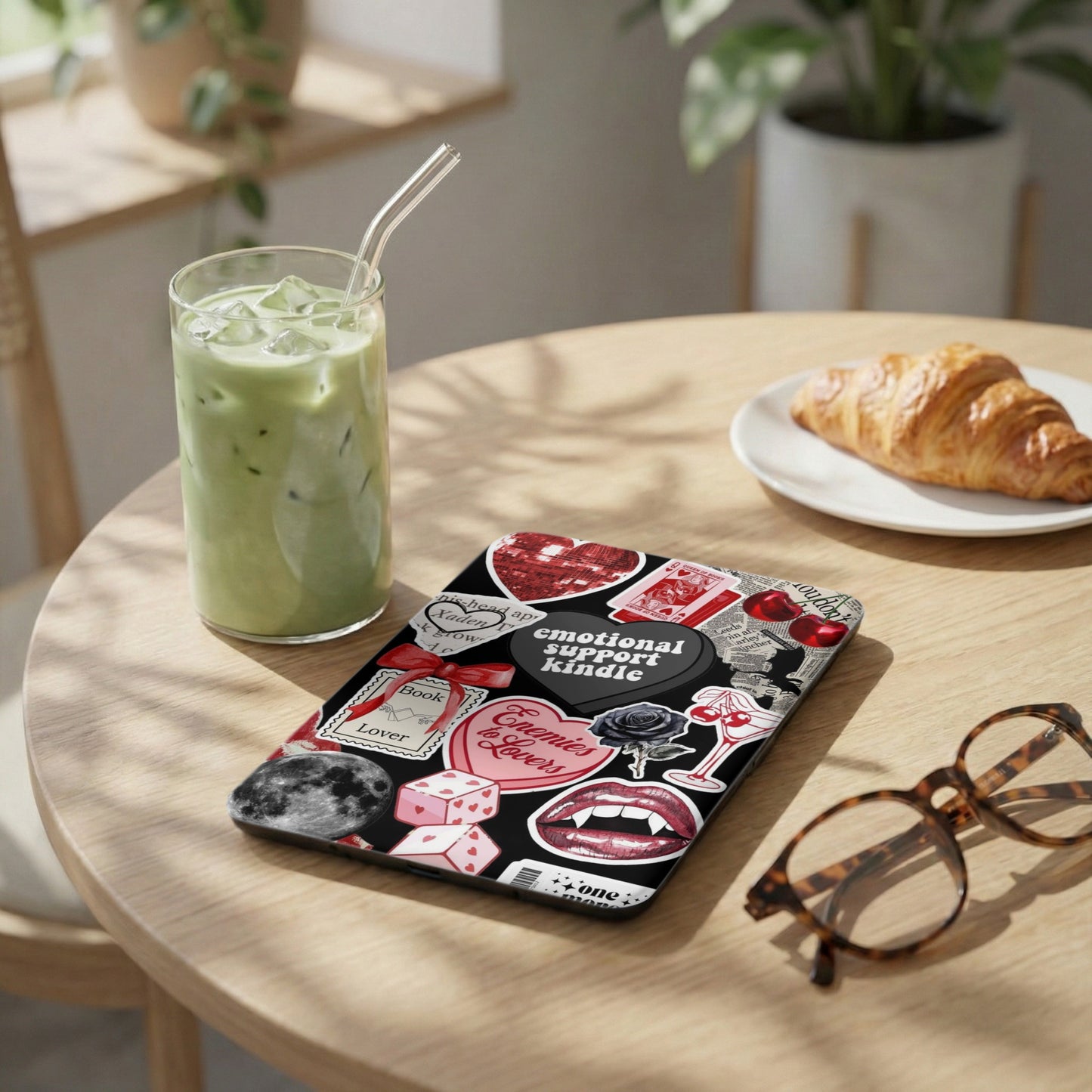 Case with "emotional support kindle" and gothic stickers next to matcha drink.