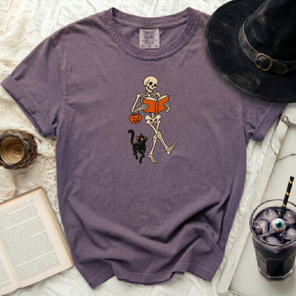 Skeleton reading book with pumpkin bucket and black cat.
