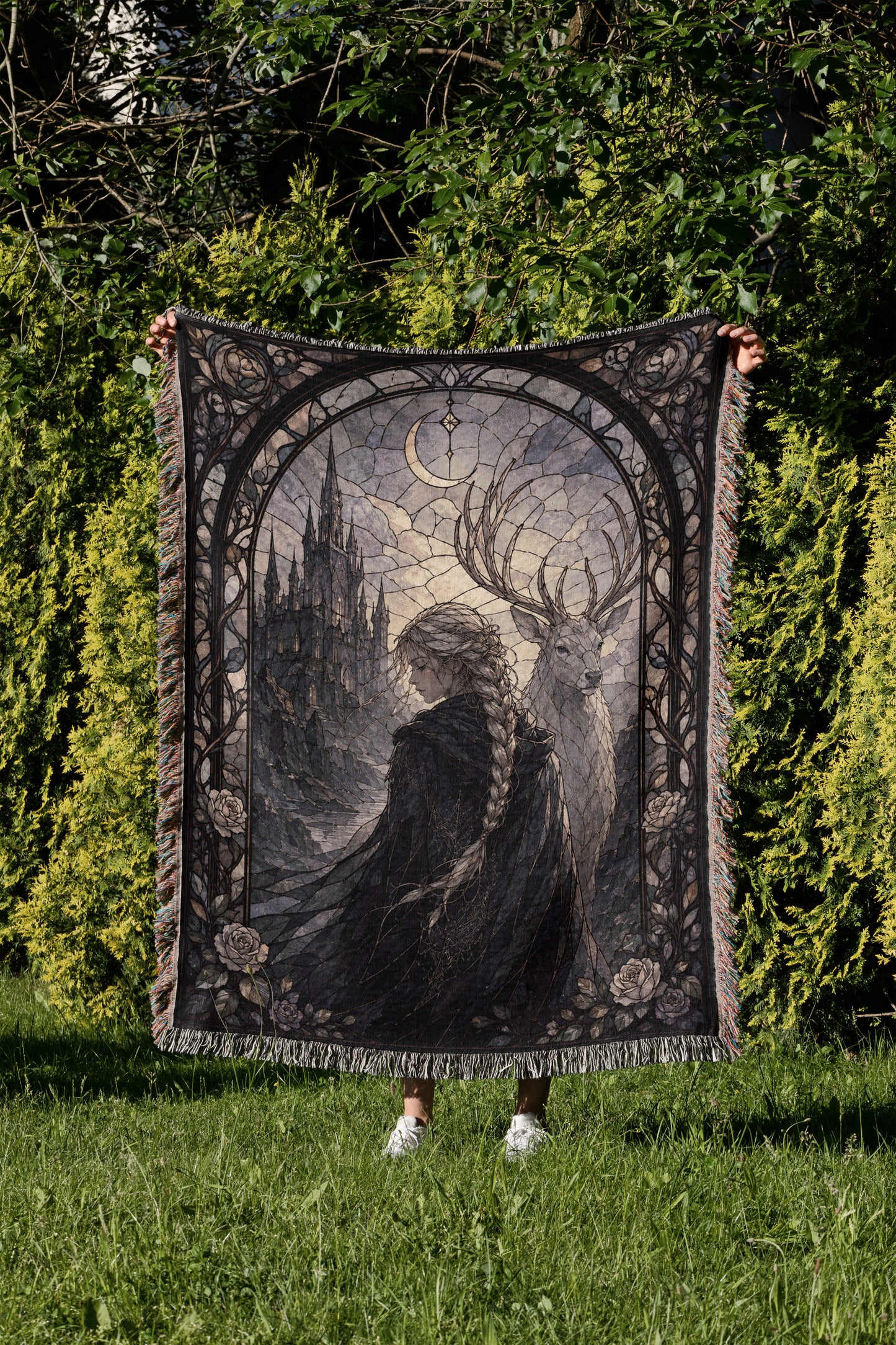 A person is holding up a large, ornate tapestry with a castle and deer design on it, set against a backdrop of trees and greenery.