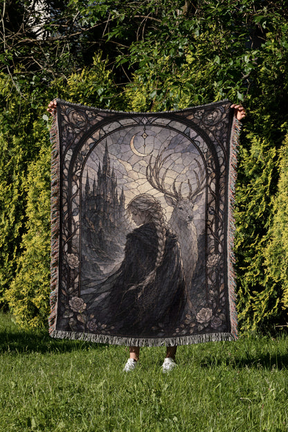 A person is holding up a large, ornate tapestry with a castle and deer design on it, set against a backdrop of trees and greenery.