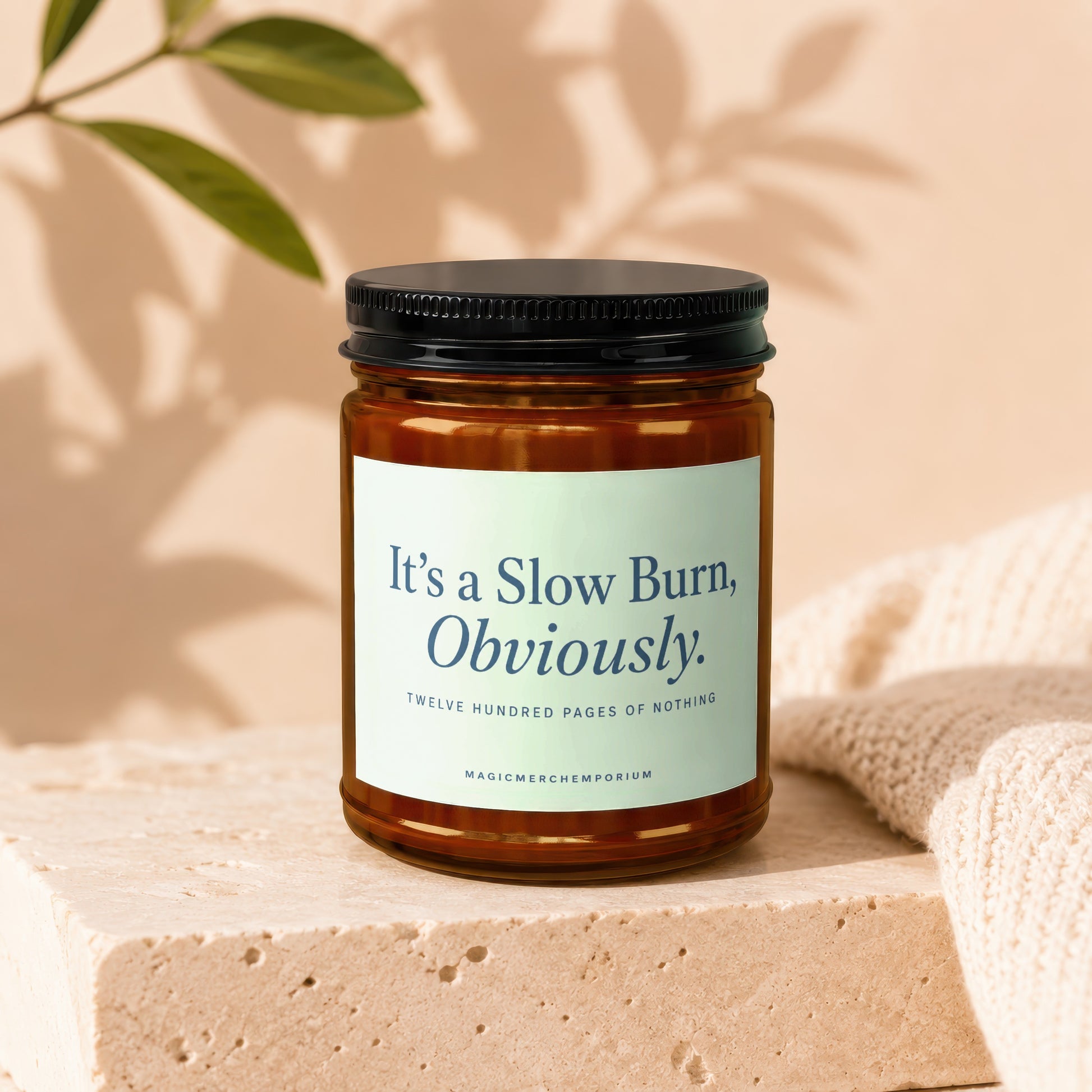 Amber jar candle with black lid labeled "It's a Slow Burn, Obviously. Twelve Hundred Pages of Nothing."