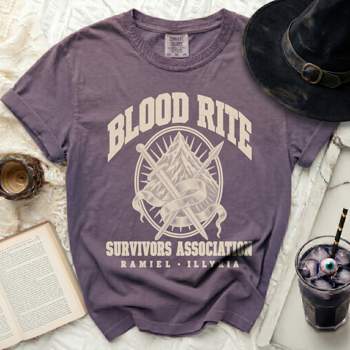 A purple t-shirt featuring the text "BLOOD RITE SURVIVORS ASSOCIATION RAMIEL ILLYRIA" surrounding a mountain, sword, and spear design.