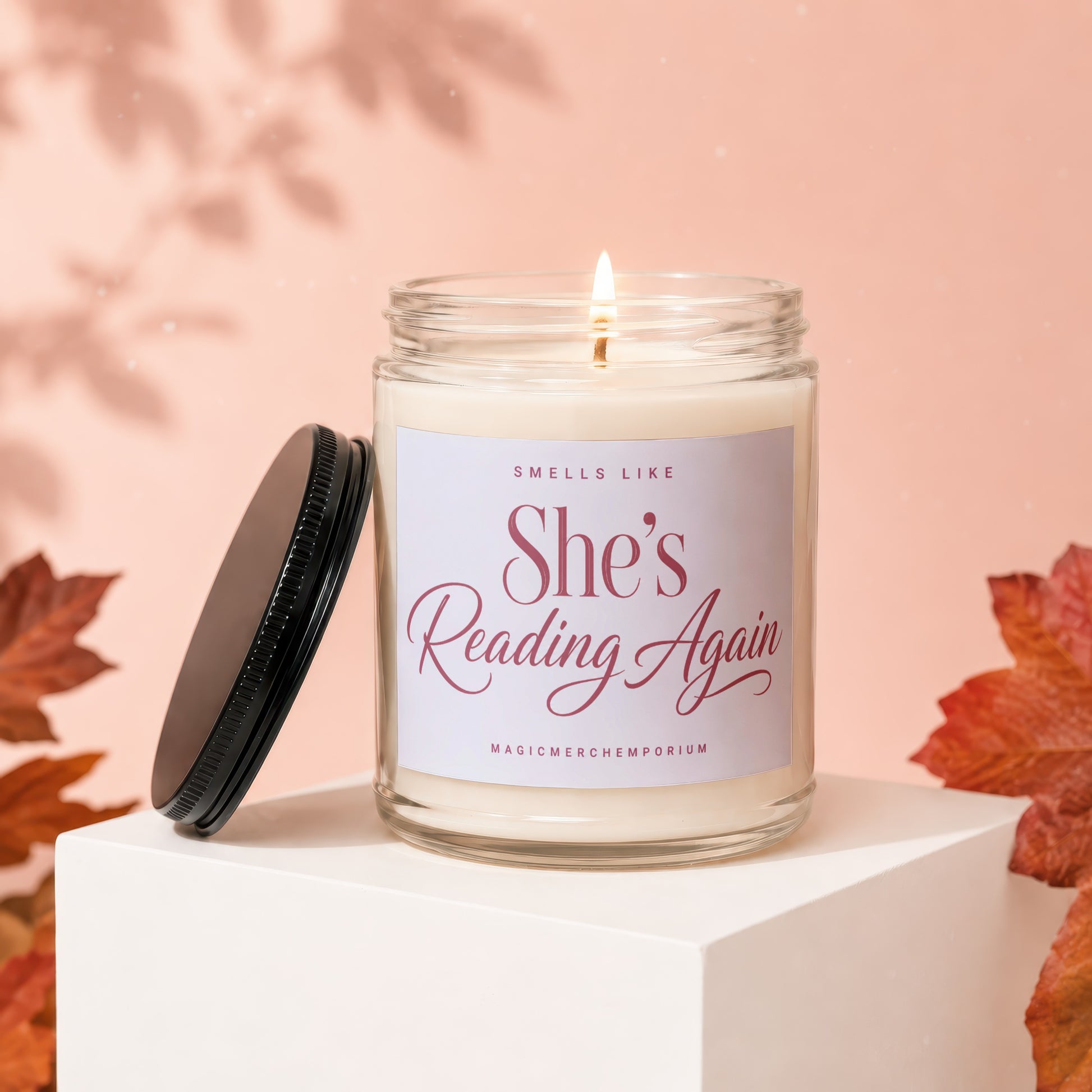 Burning candle with "She's Reading Again" text on label, with lid and leaves.