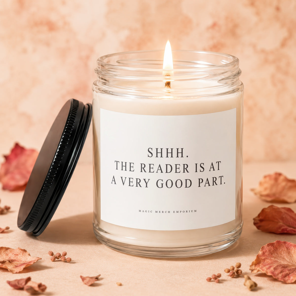 Glass candle with lit wick and black lid, reads "SHHH. THE READER IS AT A VERY GOOD PART."