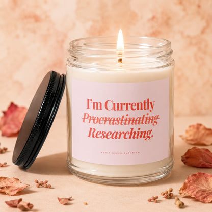 Jar candle with lit wick, pink label reads "I'm Currently Procrastinating Researching."