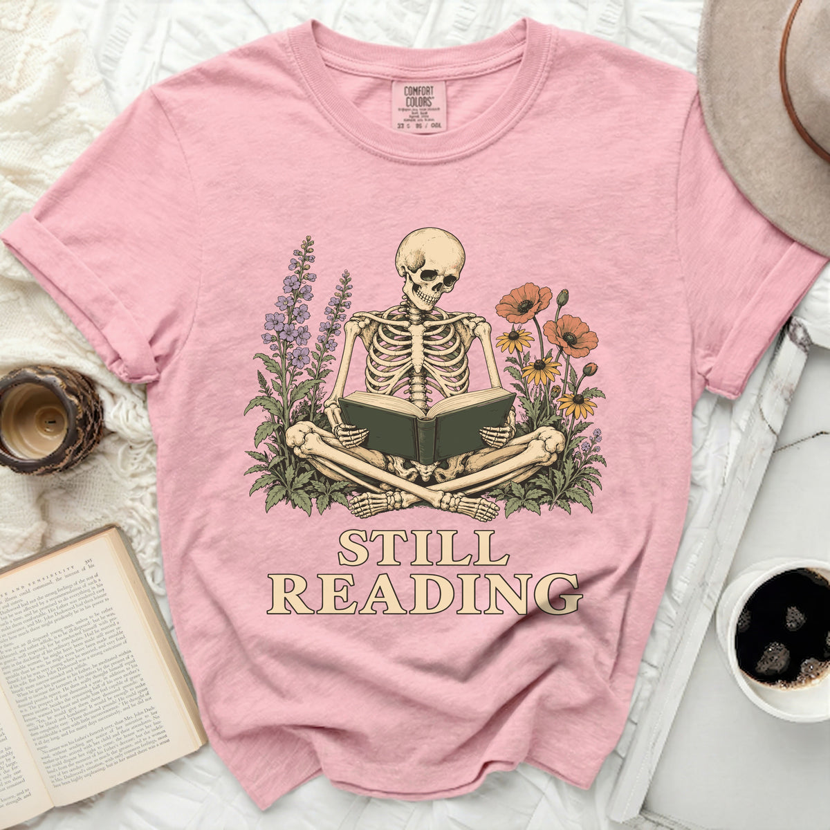 Pink t-shirt with skeleton reading a book amidst flowers, text "STILL READING".