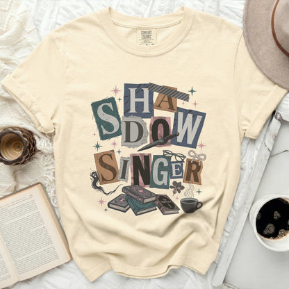 Cream t-shirt with "Shadow Singer" graphic, books, and coffee.