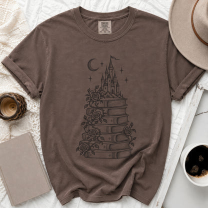 A brown t-shirt featuring a graphic print of a castle atop stacked books with climbing roses and a crescent moon.
