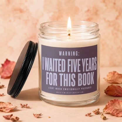 A lit glass candle with a black lid and a label reading Warning: I Waited Five Years For This Book.