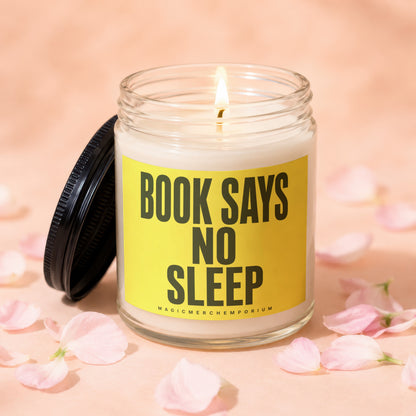 Lit candle in glass jar with yellow label reading "BOOK SAYS NO SLEEP" among pink petals.