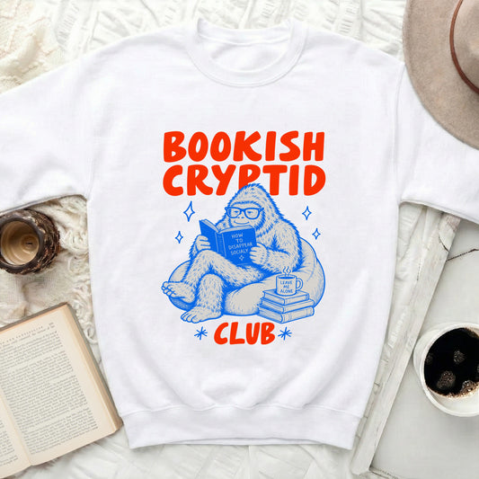 White sweatshirt with orange "Bookish Cryptid Club" graphic of a reading yeti.