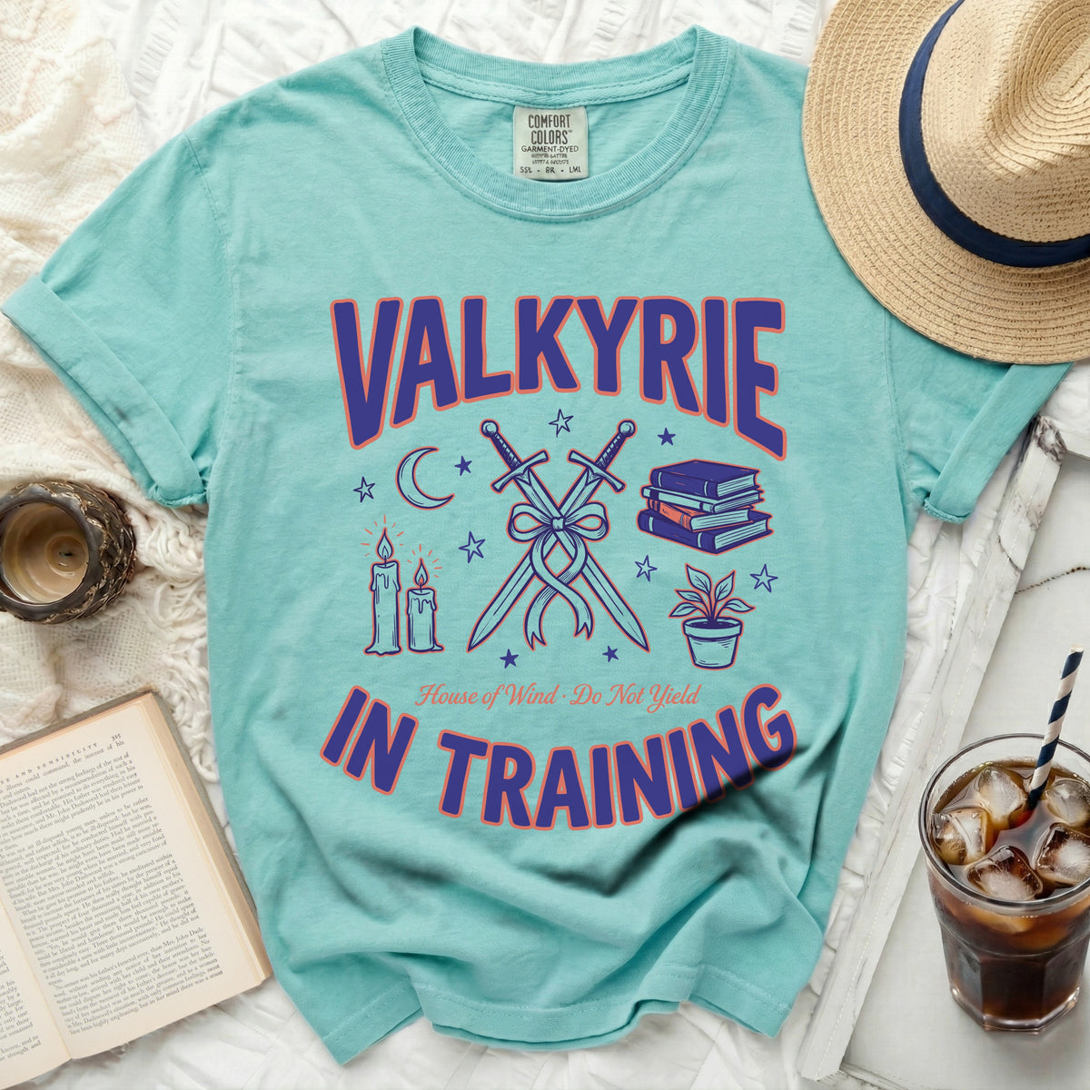 Teal t-shirt with purple and orange text reading VALKYRIE IN TRAINING and crossed swords, surrounded by books, candles, and a potted plant.