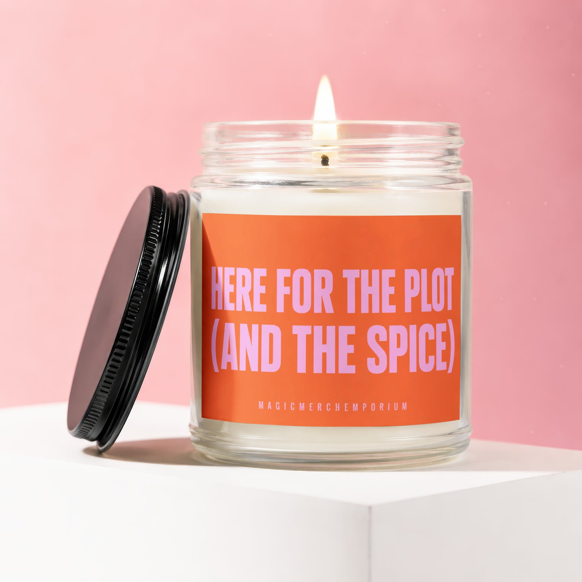 Lit candle in glass jar with orange label reading "HERE FOR THE PLOT (AND THE SPICE)".