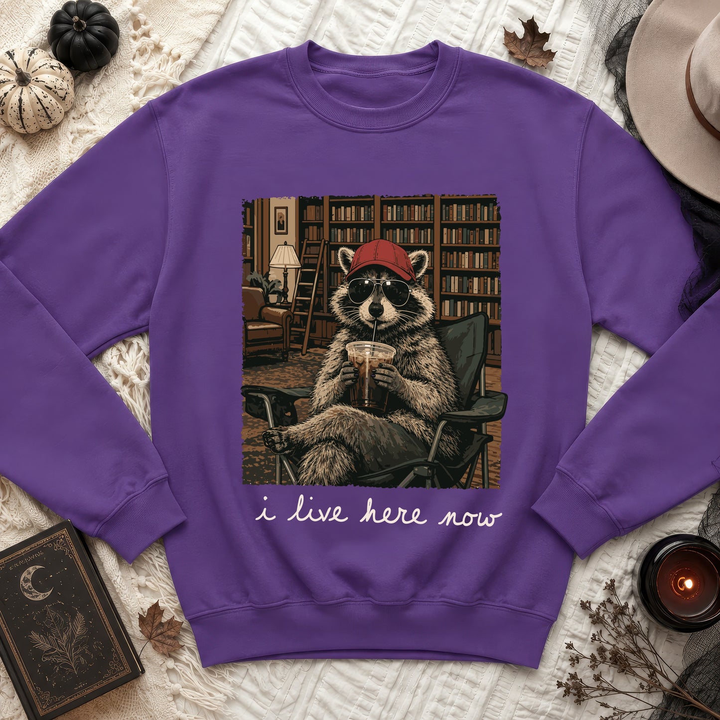 Purple sweatshirt featuring a raccoon wearing sunglasses and a cap, with text "i live here now."