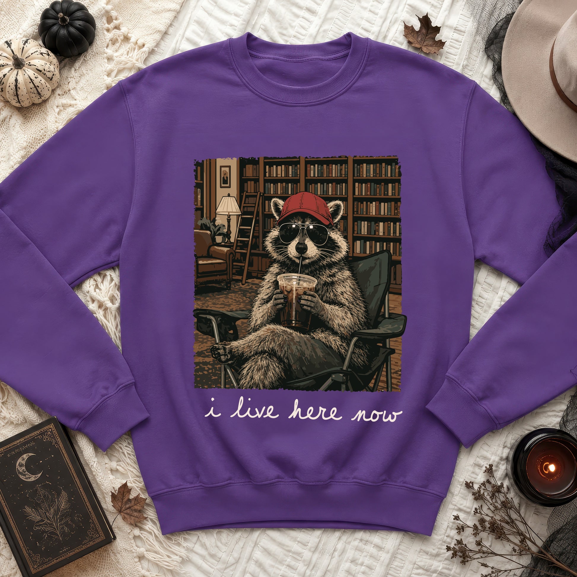 Purple sweatshirt featuring a raccoon wearing sunglasses and a cap, with text "i live here now."