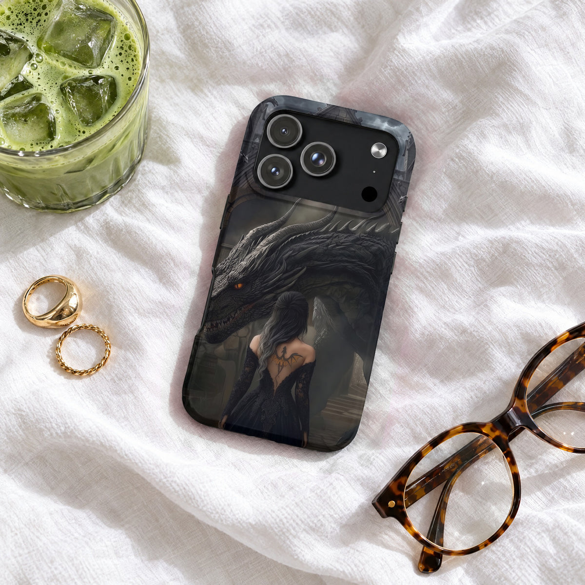 A black phone case featuring a woman and a dragon resting on a white fabric surface with glasses, rings, and an iced drink.