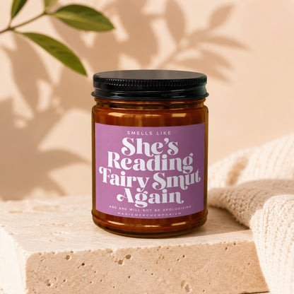 Amber jar candle with purple label reading "Smells Like She's Reading Fairy Smut Again".