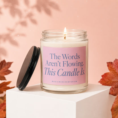 Clear glass jar candle with pink label and blue text, lit wick.