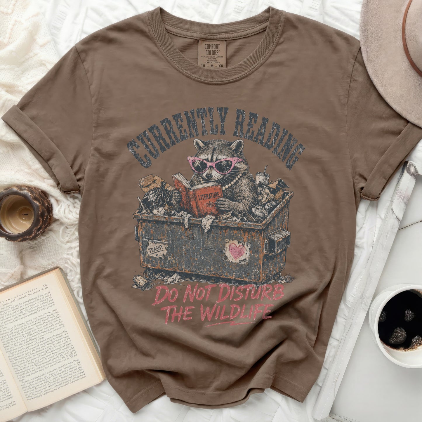 T-shirt with a raccoon reading in a trash can graphic and text.