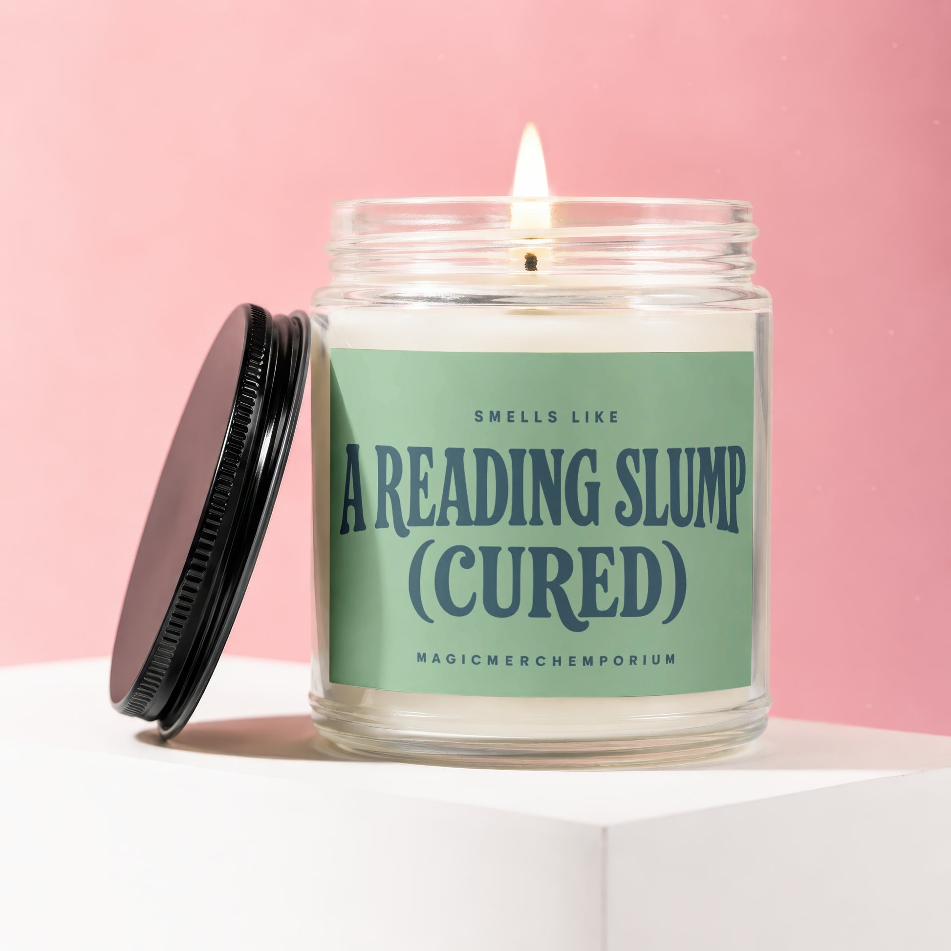 Candle in a jar with a lit wick and a green label reading "Smells Like A Reading Slump (Cured)".