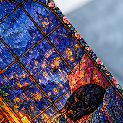 A black cat sleeps curled up beside a stained glass window depicting a city at night.