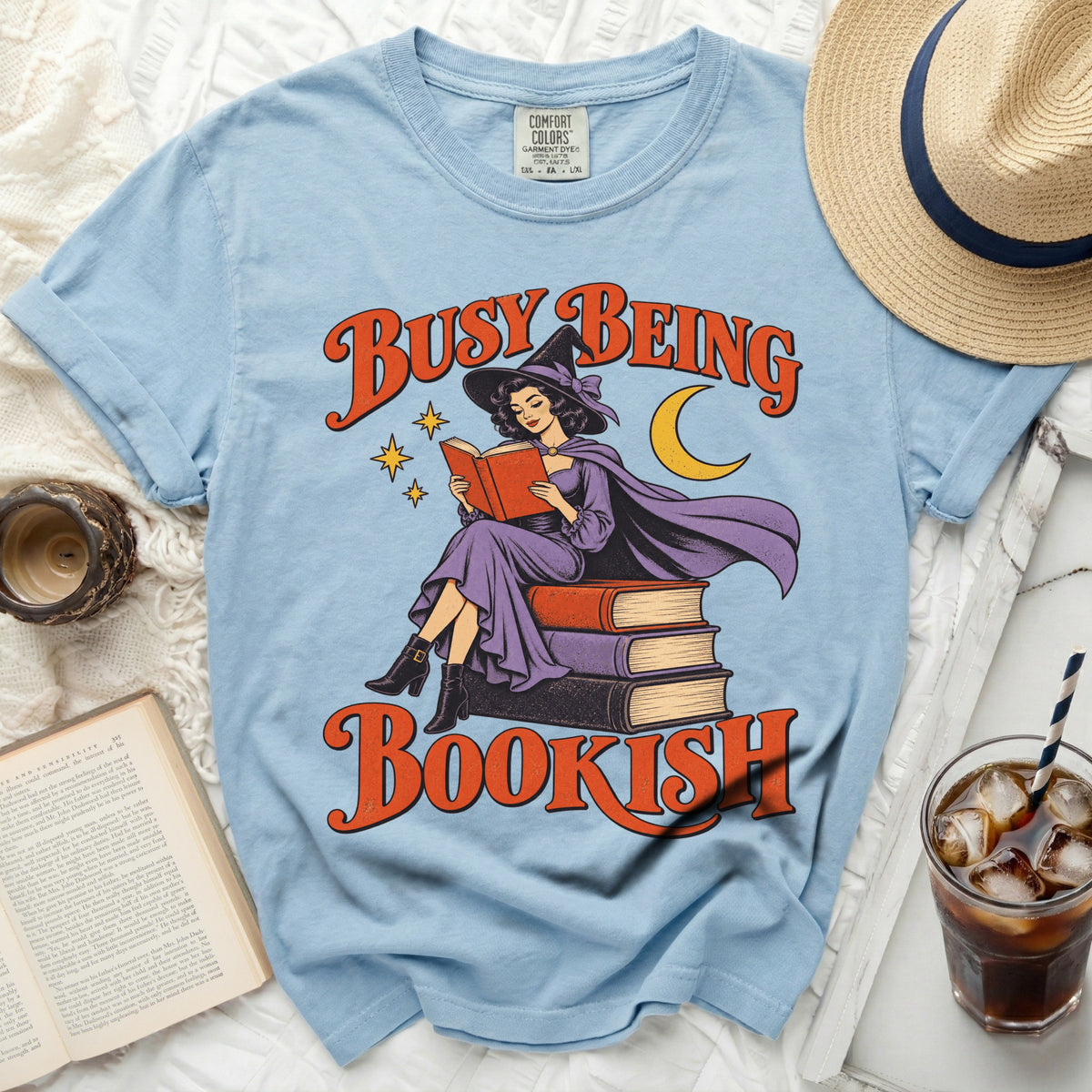 Light blue t-shirt with witch reading books and text "BUSY BEING BOOKISH".