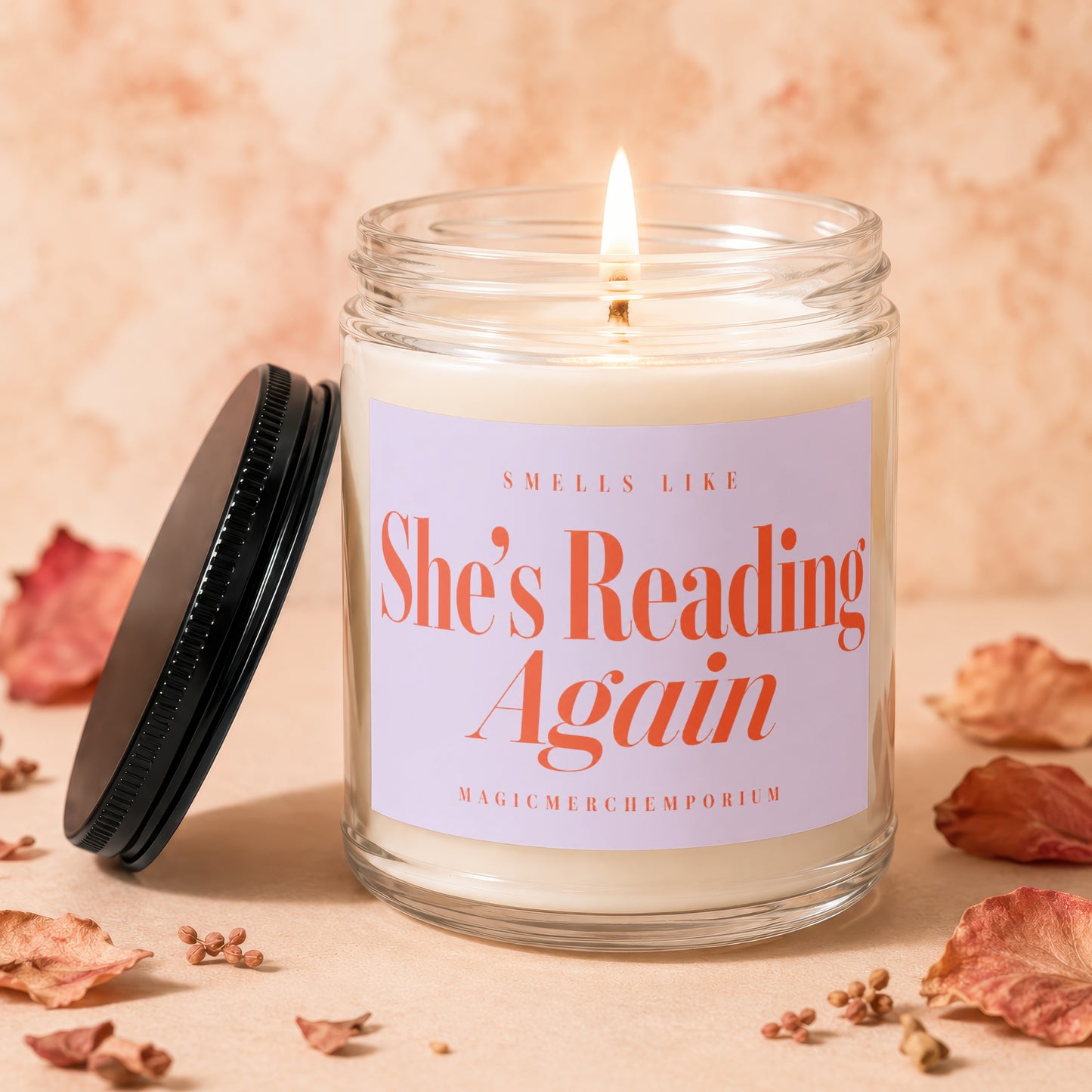 Scented candle with "She's Reading Again" on label, lit flame, black lid.