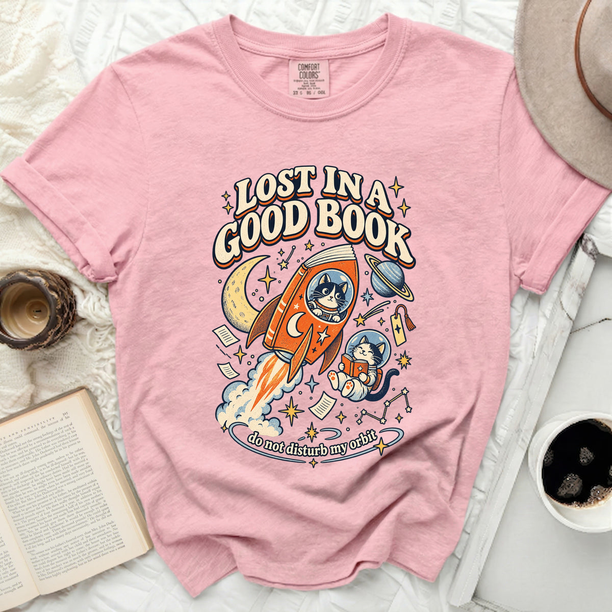 Pink t-shirt with graphic of cats, rocket, moon, stars, and text "LOST IN A GOOD BOOK".