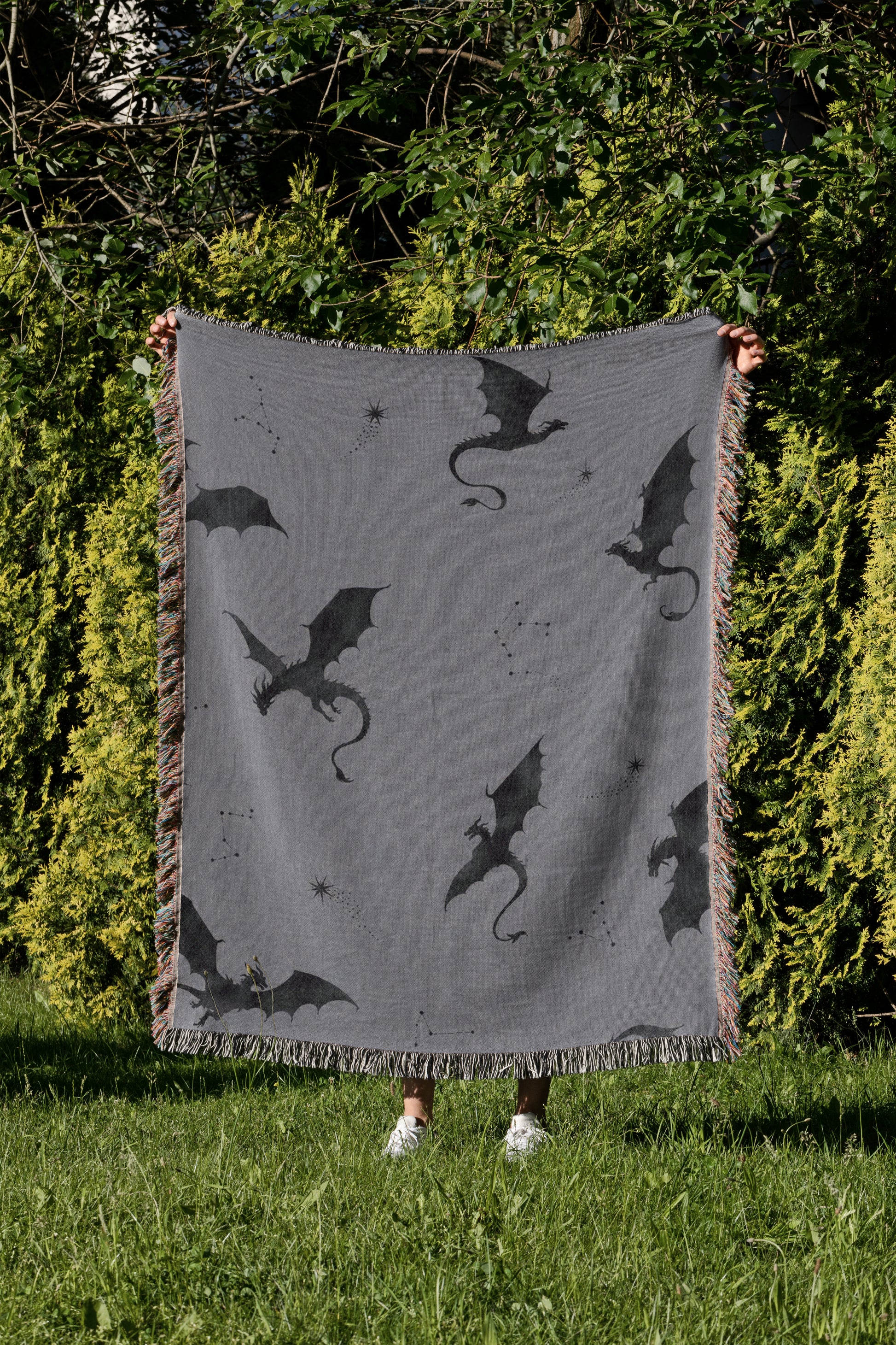 A person is holding up a gray blanket with black dragon silhouettes on it, standing in a grassy area with trees in the background.