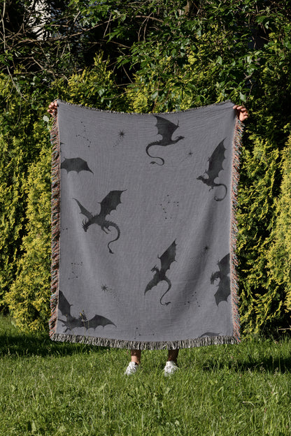 A person is holding up a gray blanket with black dragon silhouettes on it, standing in a grassy area with trees in the background.