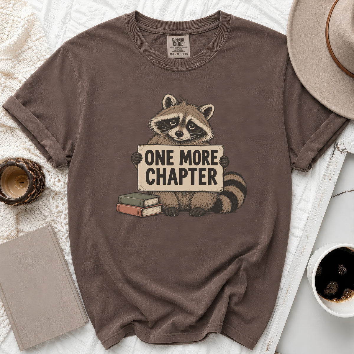 Brown t-shirt with graphic of raccoon holding sign "ONE MORE CHAPTER" and books.