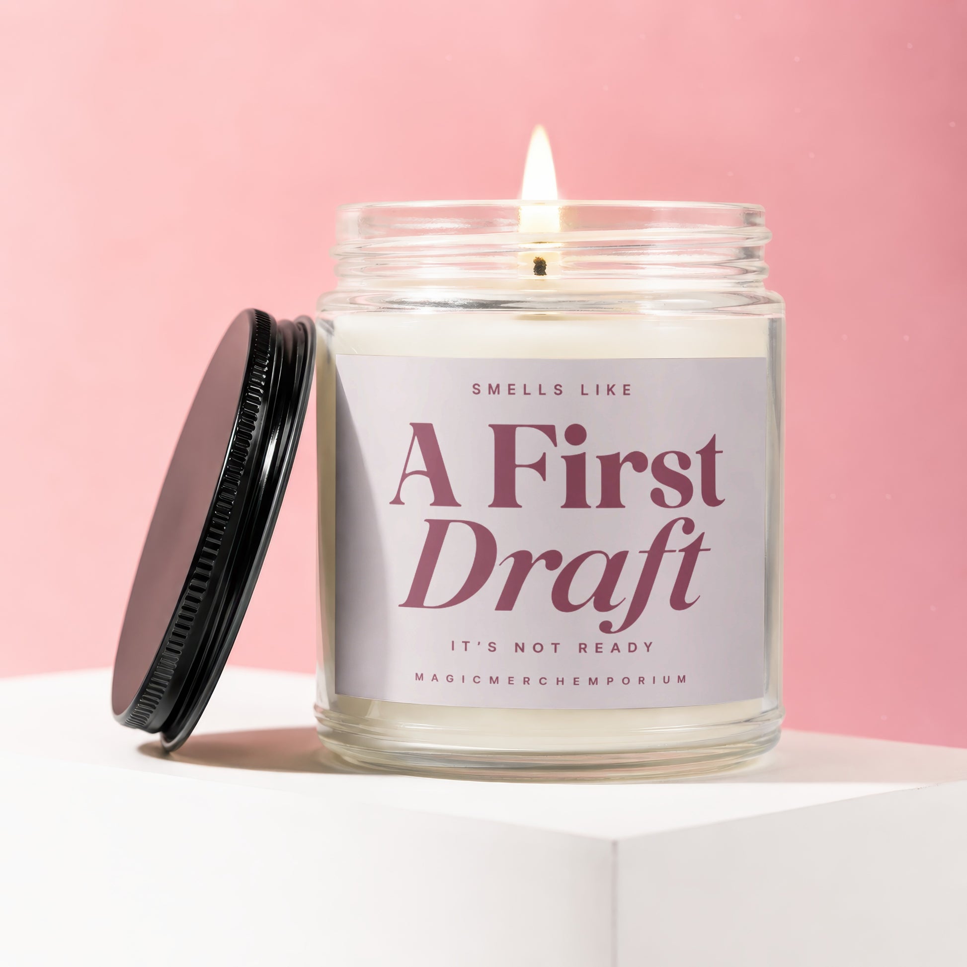 Lit candle in a glass jar with "SMELLS LIKE A FIRST DRAFT IT'S NOT READY" label.