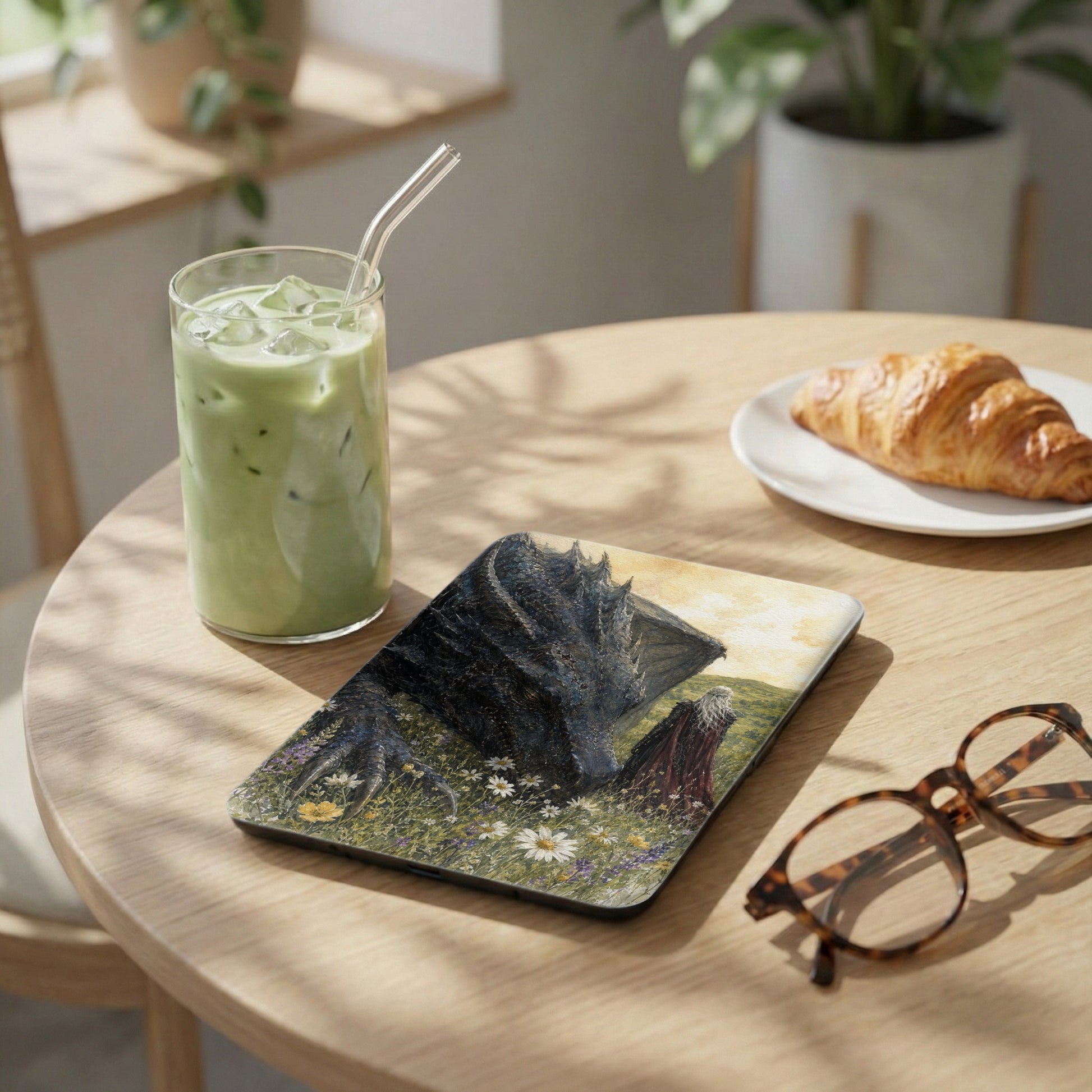Tablet with a dragon and person in a field, next to iced matcha tea and croissant.