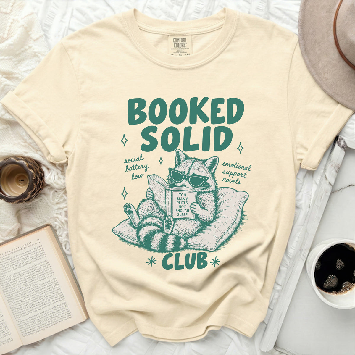 Booked Solid Club t-shirt with raccoon reading a book, "Too Many Plots, Not Enough Sleep".