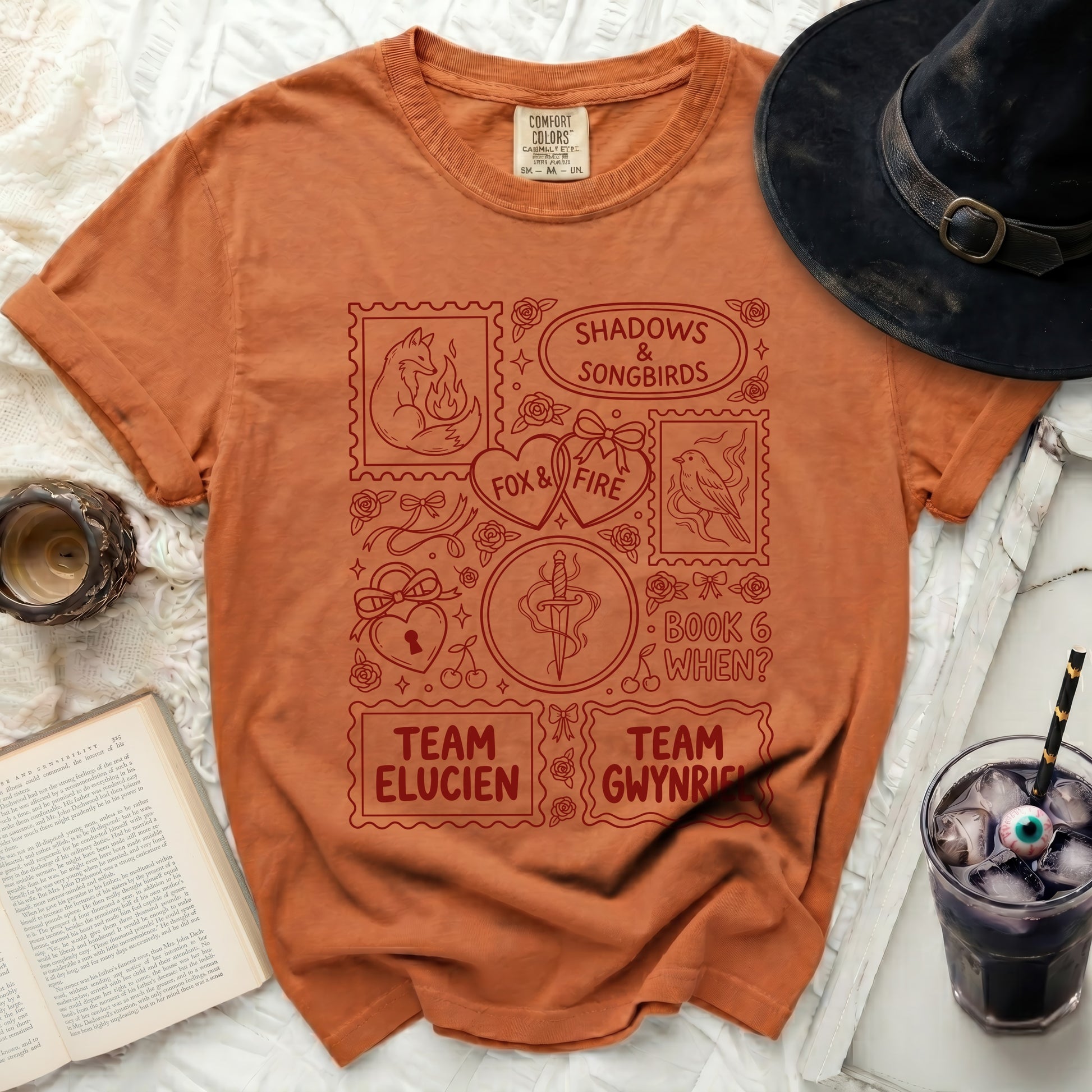 Orange t-shirt with red graphic featuring stamps with foxes, birds, hearts, sword, and text.