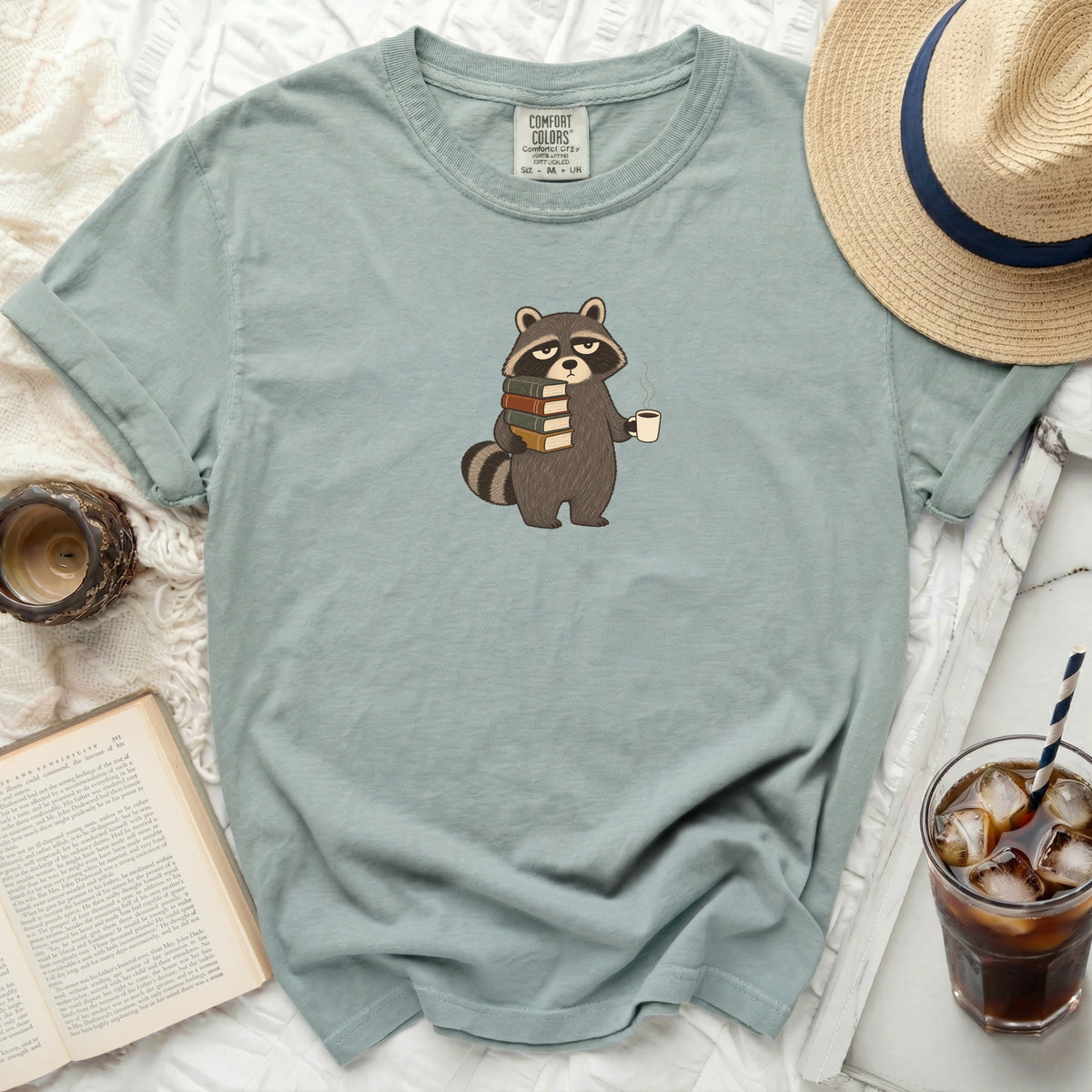 Light blue t-shirt with a graphic of a raccoon holding books and coffee.