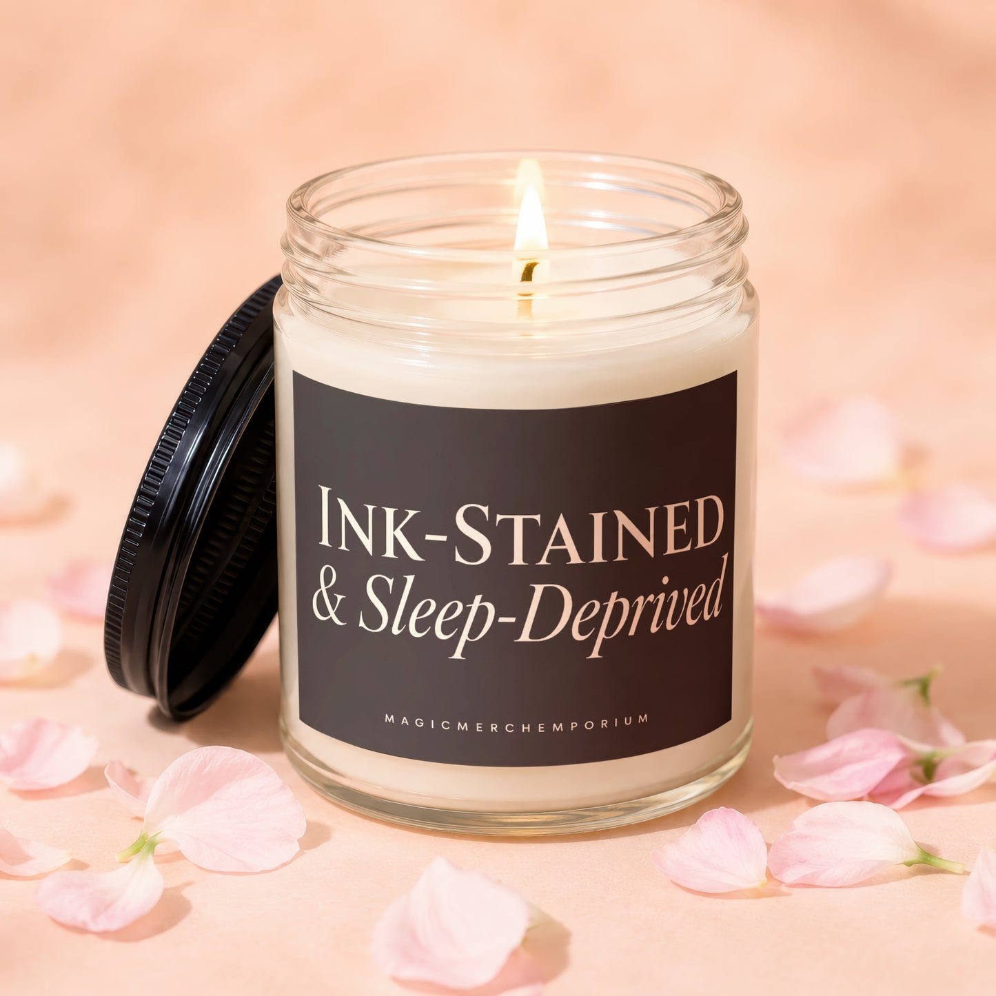 Glass candle burning with "Ink-Stained & Sleep-Deprived" label and pink petals.