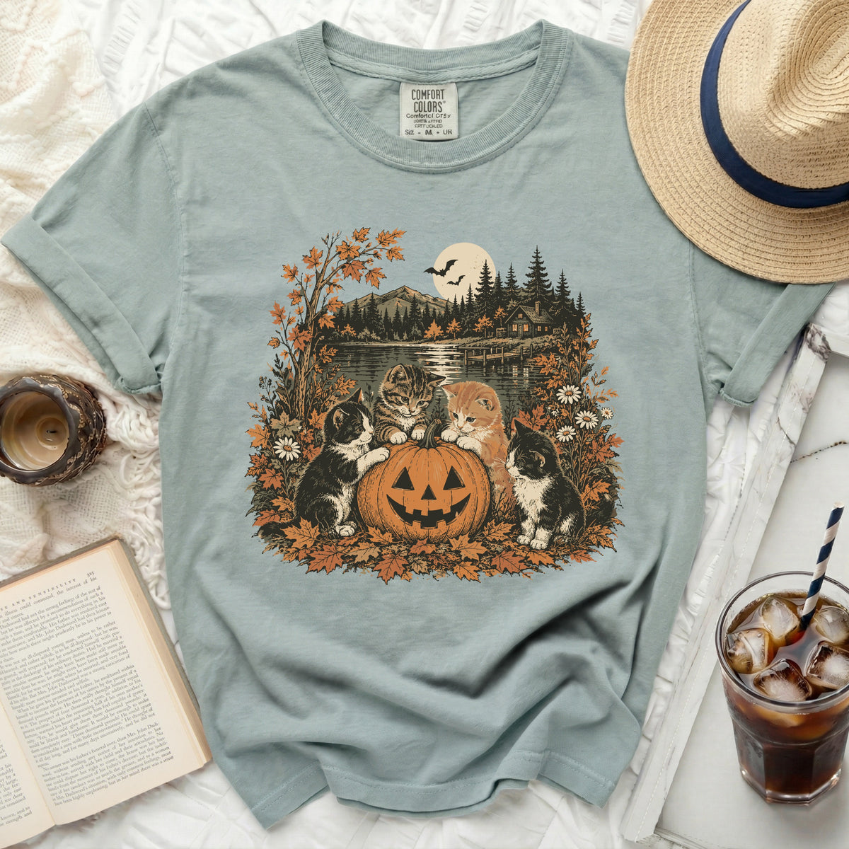 Four kittens sit near a jack-o'-lantern with a fall landscape on a t-shirt.