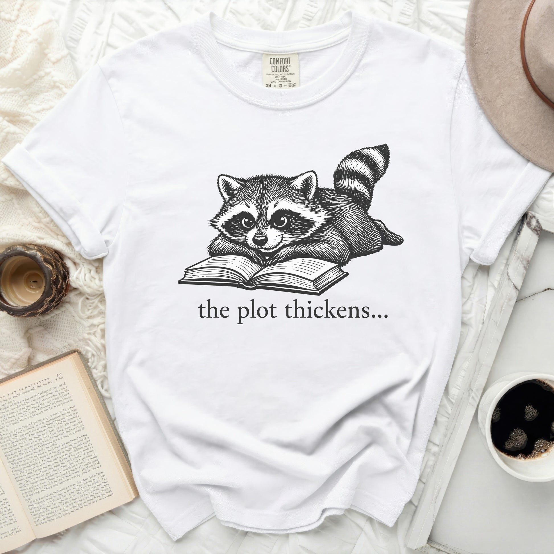 White t-shirt with a drawing of a raccoon reading a book and the text "the plot thickens..."