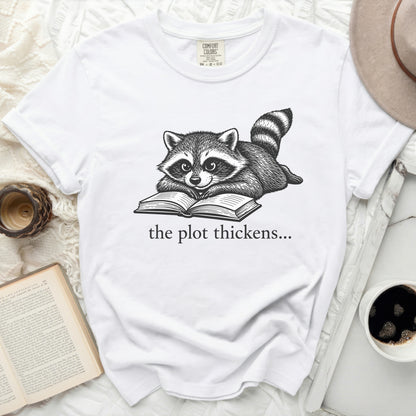 White t-shirt with a drawing of a raccoon reading a book and the text "the plot thickens..."