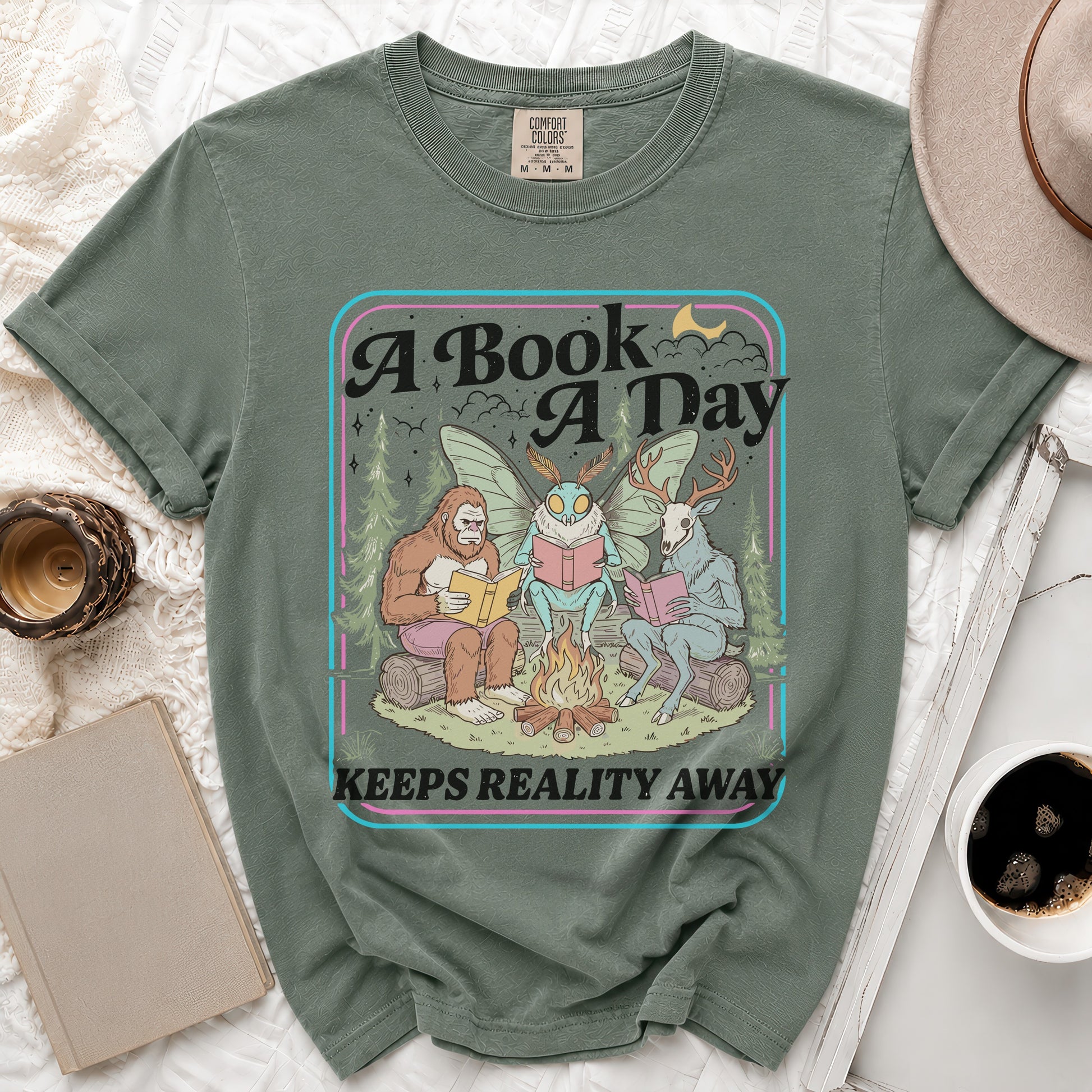 Green t-shirt with Bigfoot, mothman, and deer reading by a campfire; "A Book A Day Keeps Reality Away" text.