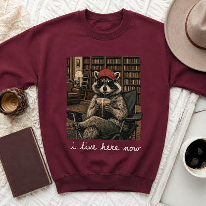 Maroon sweatshirt with a print of a raccoon wearing sunglasses and a red hat, sitting in a chair with a drink. Text below reads "i live here now."